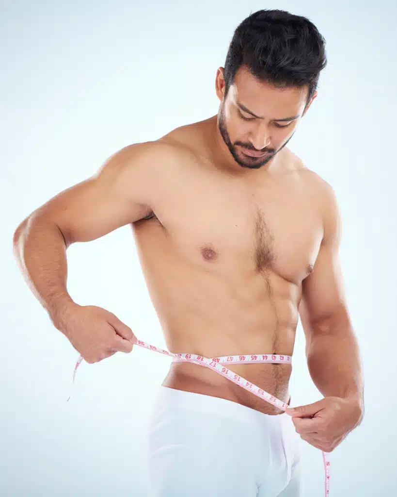 A shirtless man in white shorts measures his waist with a tape measure, focused expression, highlighting fitness and health awareness.