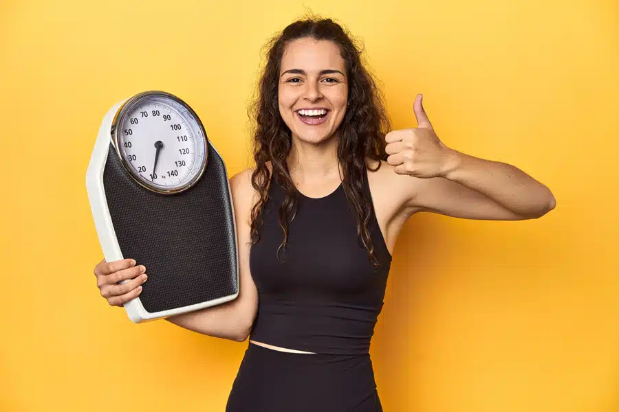 Smiling woman in a black sports outfit holds a bathroom scale and gives a thumbs-up against a vibrant yellow background, conveying positivity.