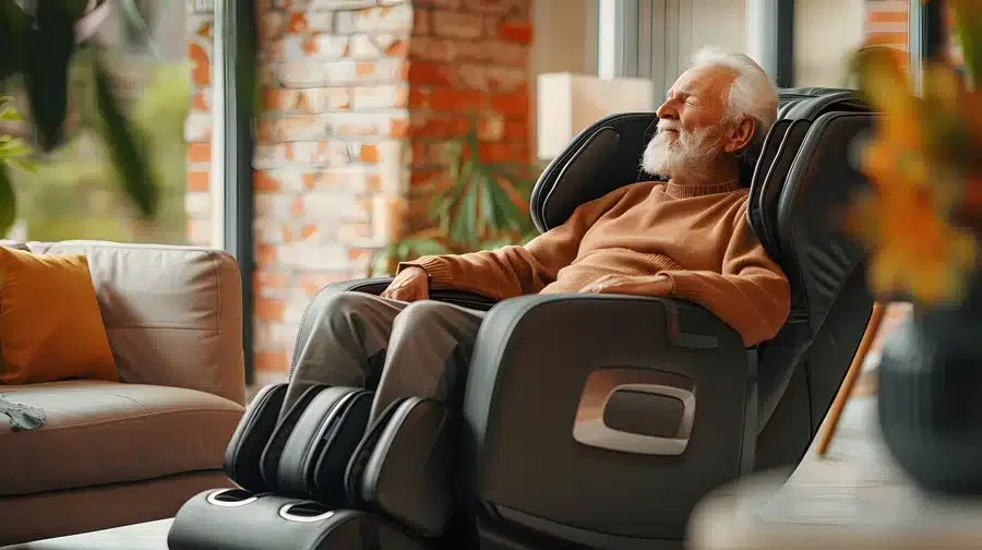 Elderly man relaxes in a massage chair at home, eyes closed and smiling. Sunlight streams through large windows, creating a warm, serene ambiance.