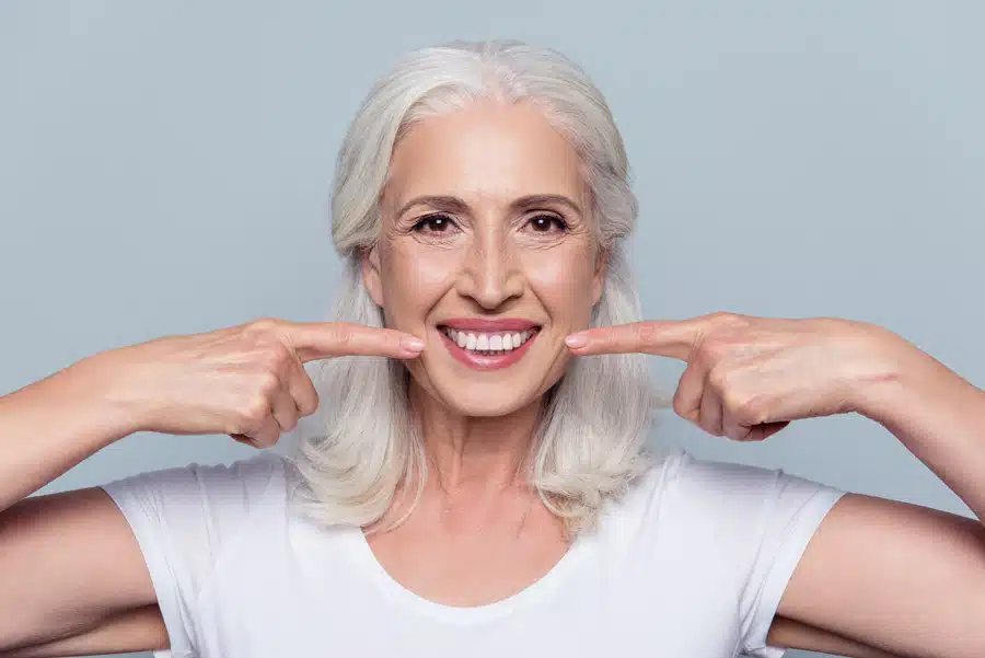 Smiling elderly woman with white hair points at her cheeks, expressing joy and positivity. She wears a white shirt, against a light grey background.