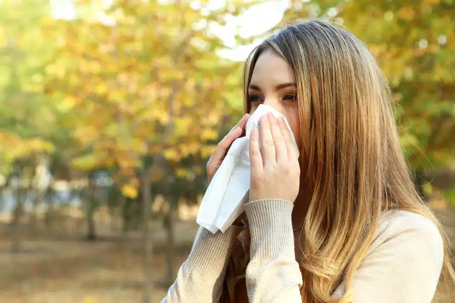 Woman with long hair sneezes into a tissue amidst a backdrop of autumnal trees. The scene conveys discomfort and suggests seasonal allergies.