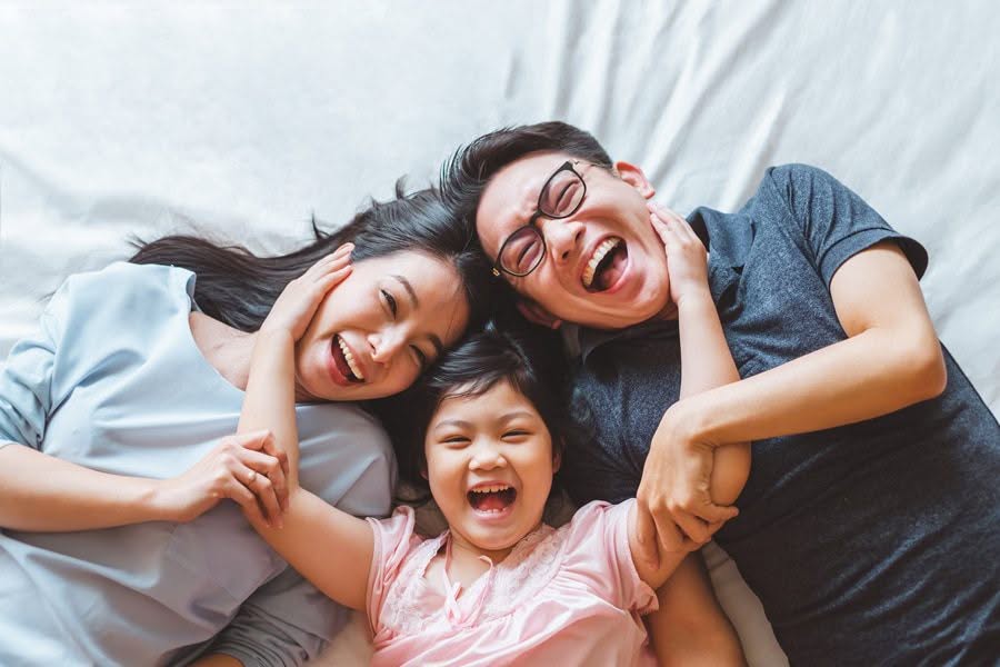 A joyful family of three lying on a bed, laughing together. They cuddle closely, expressing happiness and connection in a warm, affectionate moment.