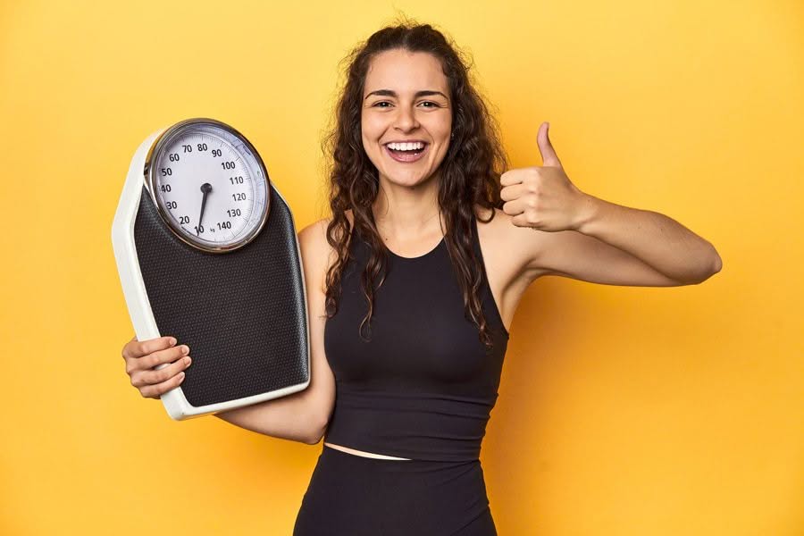 Smiling woman in a black sports outfit holds a bathroom scale and gives a thumbs-up against a vibrant yellow background, conveying positivity.