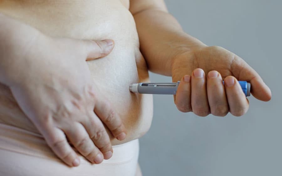 A person uses an injection pen on their abdomen. The setting is neutral, emphasizing healthcare and personal care. The mood is focused and calm.