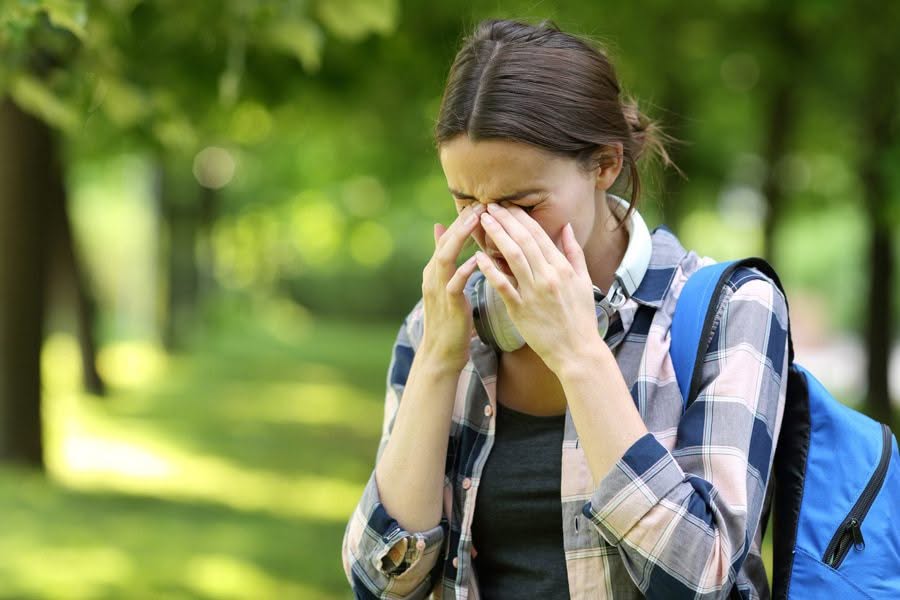 A woman outdoors in a park looks stressed, rubbing her forehead. She wears headphones around her neck and a backpack, with trees blurred in the background.