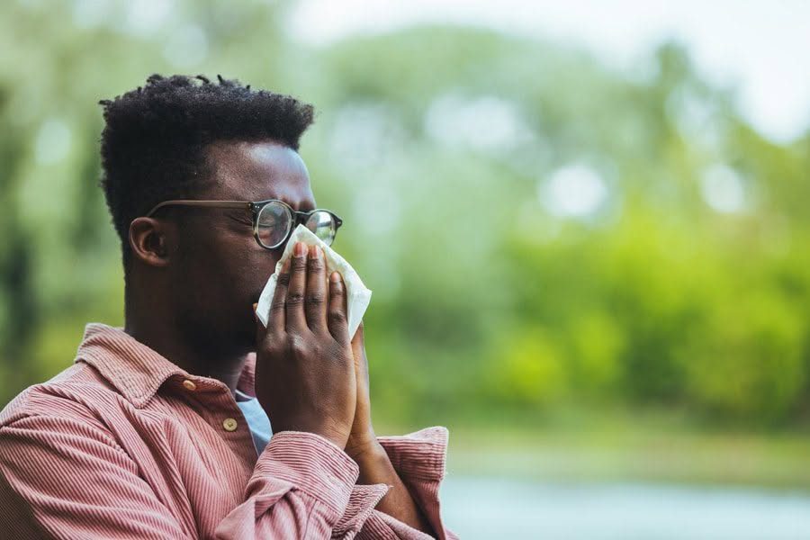 A man in glasses and a pink shirt is outdoors, using a tissue to blow his nose. The background is blurred green foliage, conveying a natural setting.